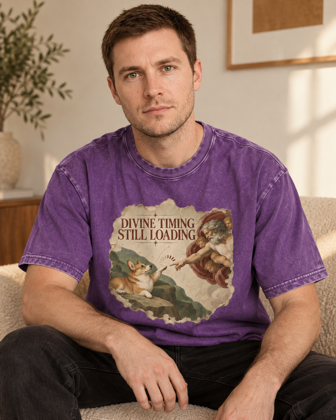 Adult model wears Purple Corgi Divine Timing Still Loading Creation Parody washed T-shirt in a seated indoor portrait