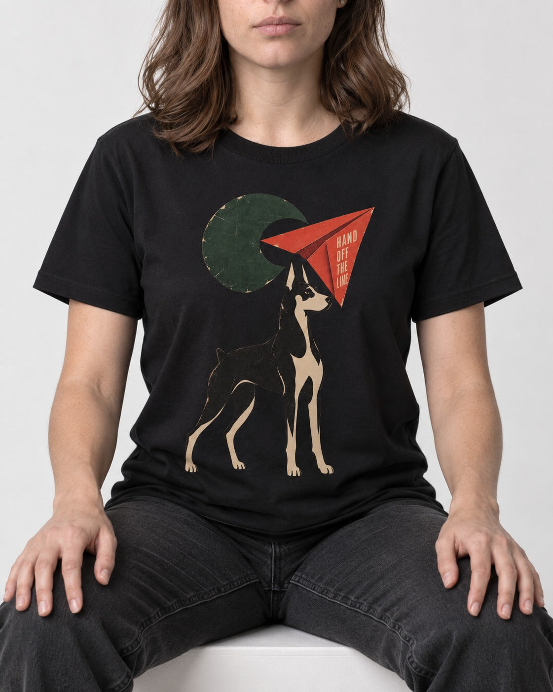 Doberman Hand Off the Line Paper Plane Graphic T-Shirt in Black, worn by a seated model