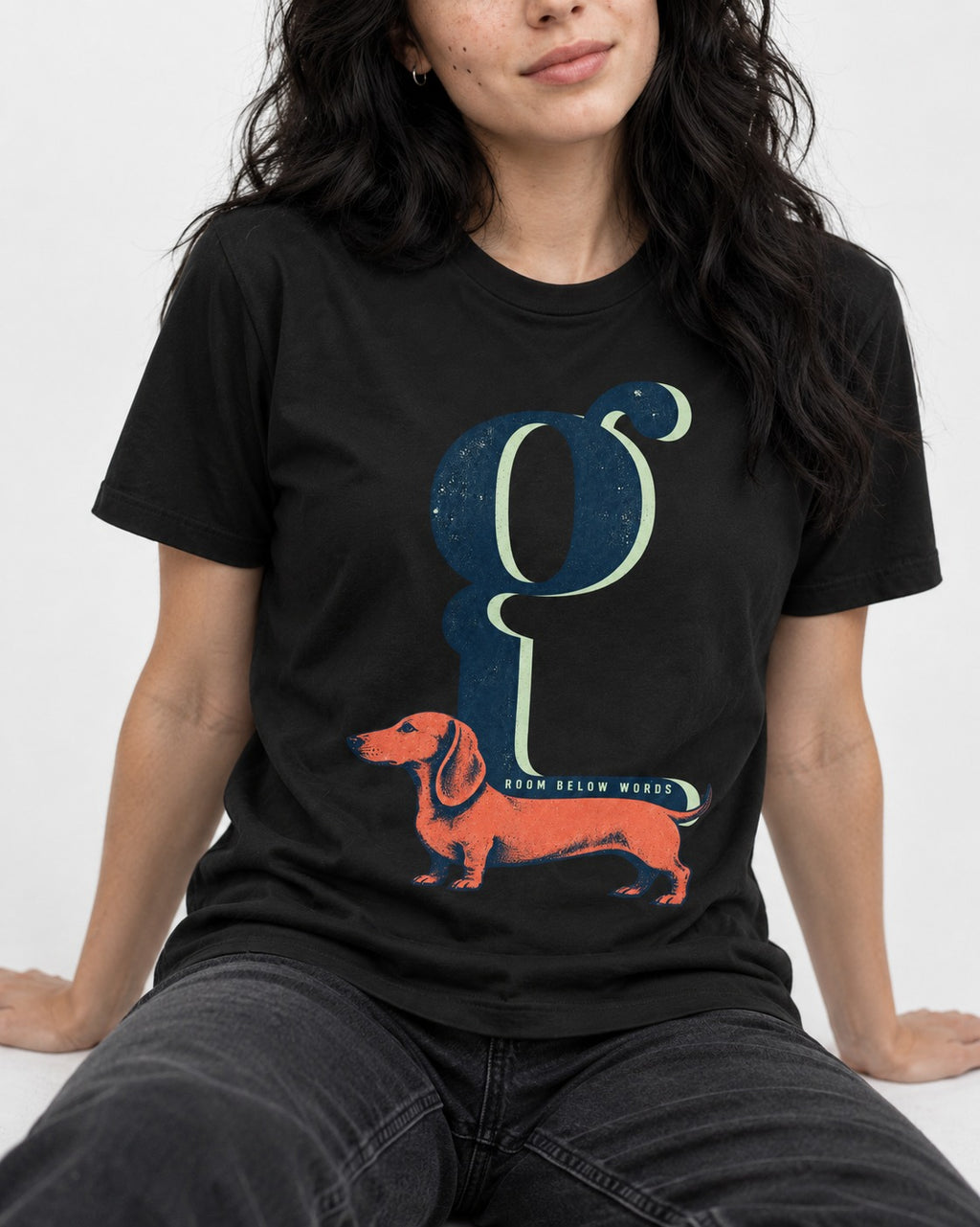 Dachshund Room Below Words Lowercase G Graphic T-Shirt in Black, worn by a seated model