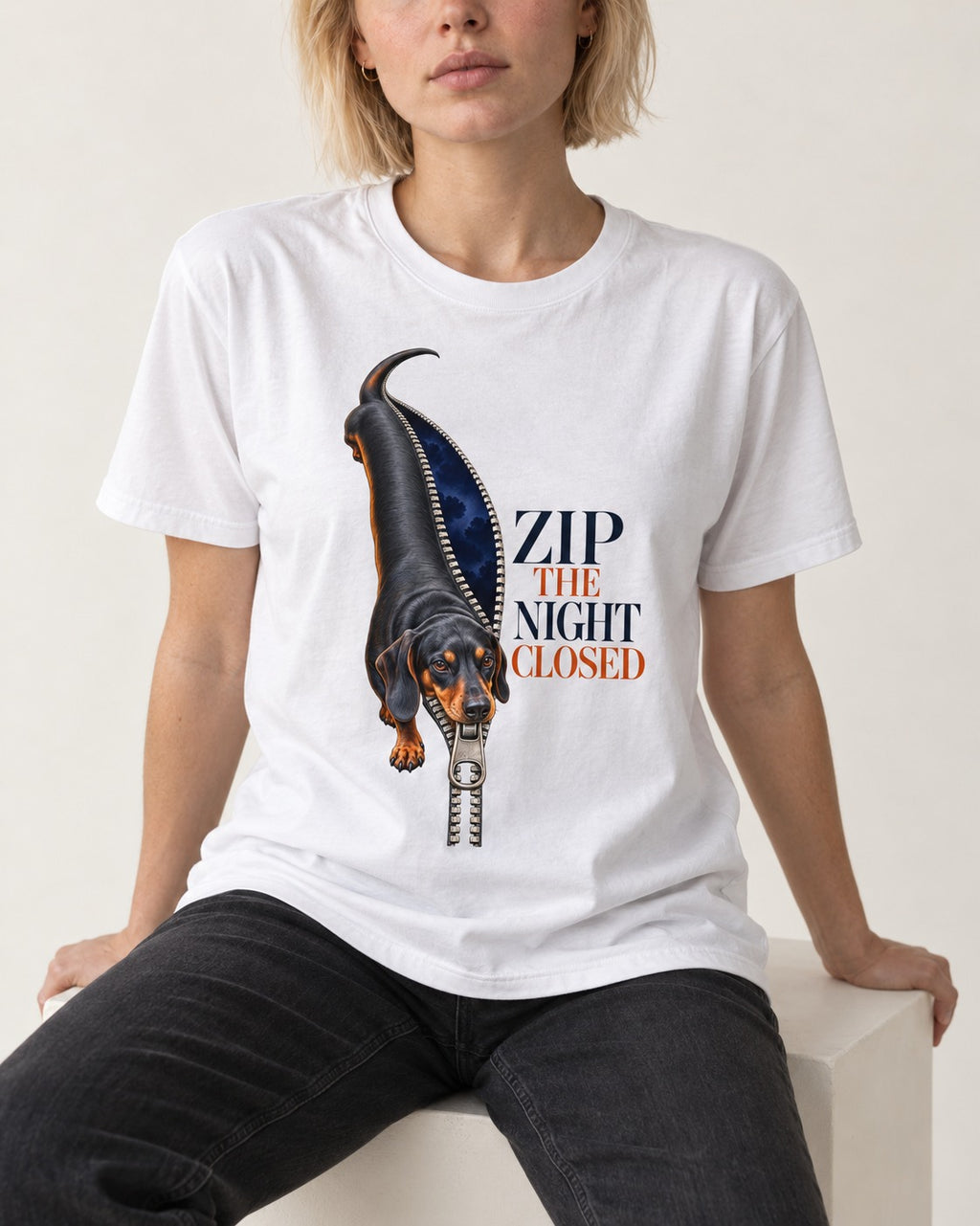 Zip the Night Closed Dachshund T-Shirt in White, worn by a seated model