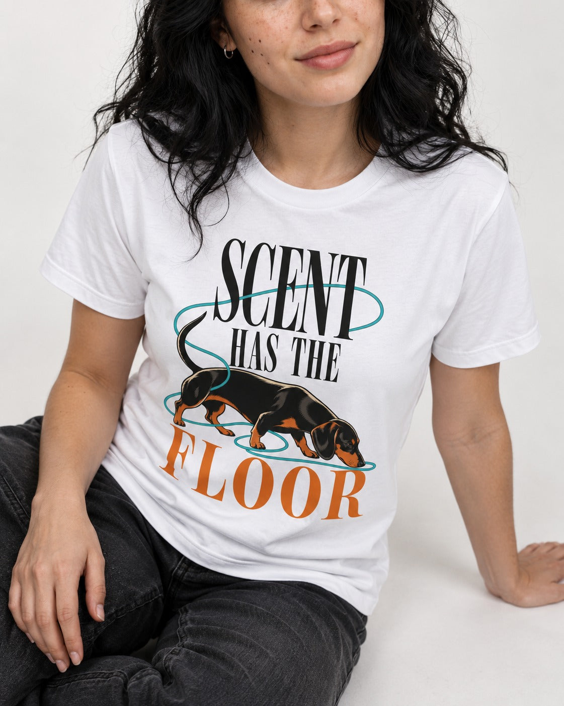 Dachshund Scent Has the Floor T-Shirt in White, worn by a seated model