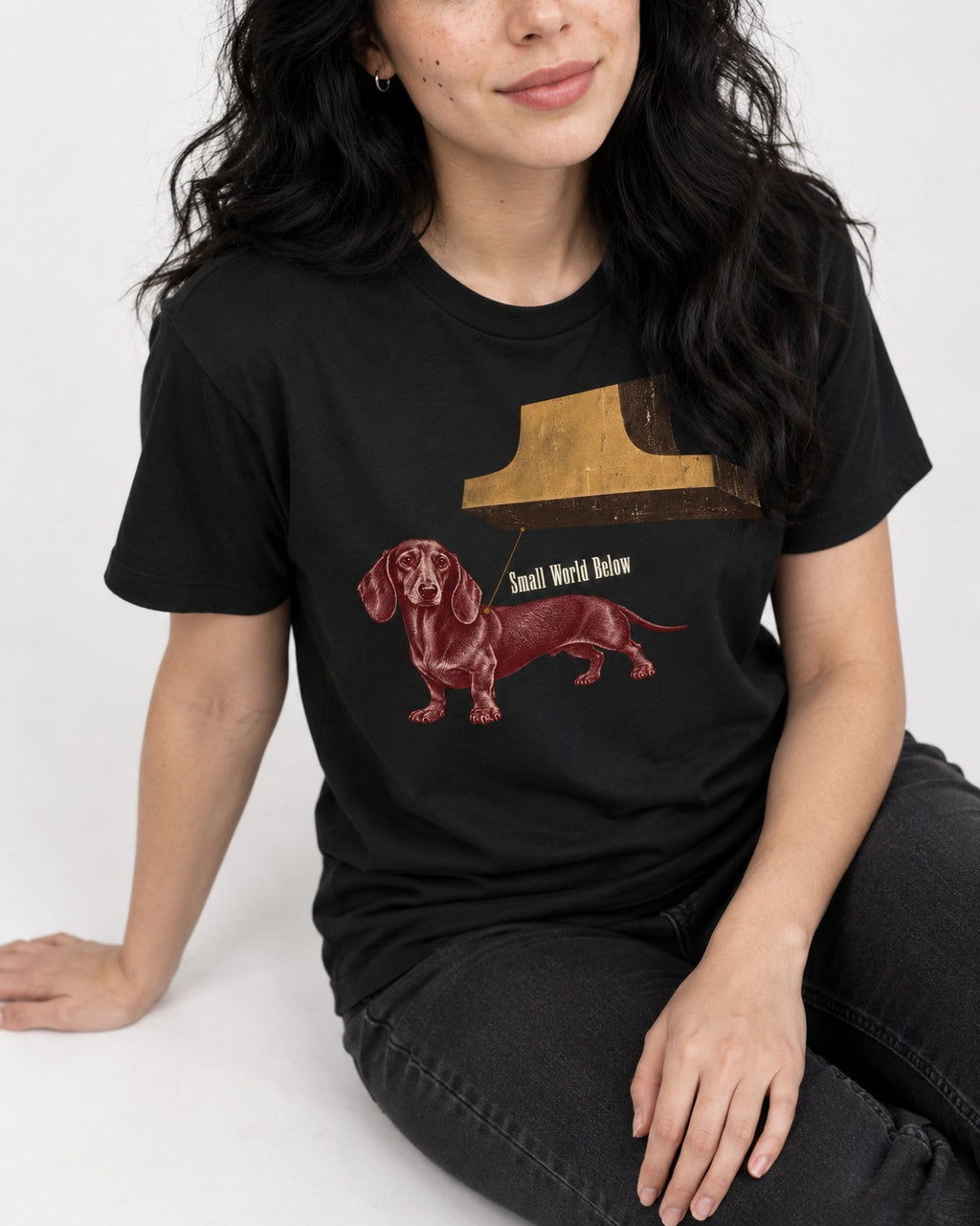 Dachshund Small World Below Giant Shoe Graphic T-Shirt in Black, worn by a seated model