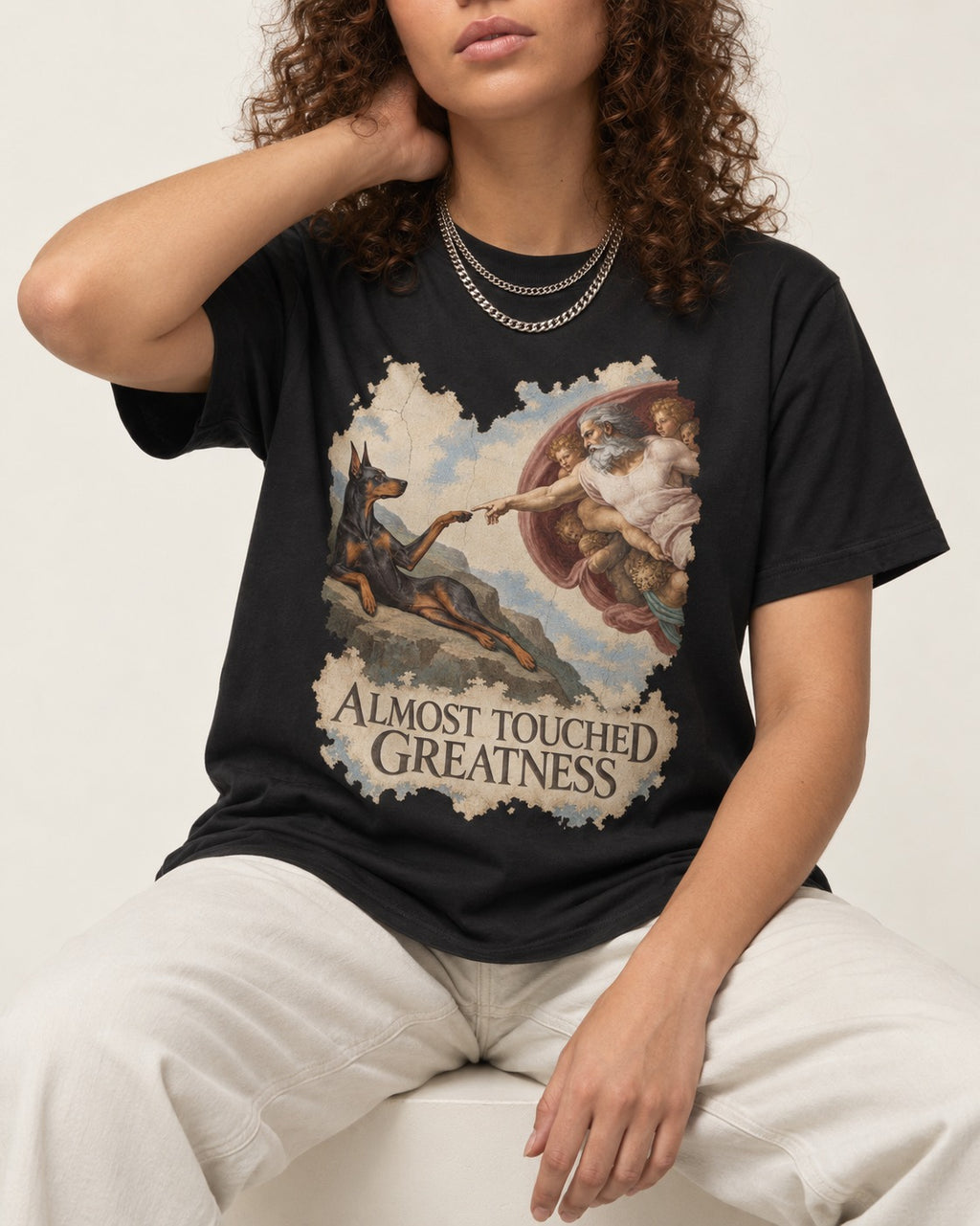 Doberman Almost Touched Greatness Creation Scene Graphic T-Shirt in Black, worn by a seated model