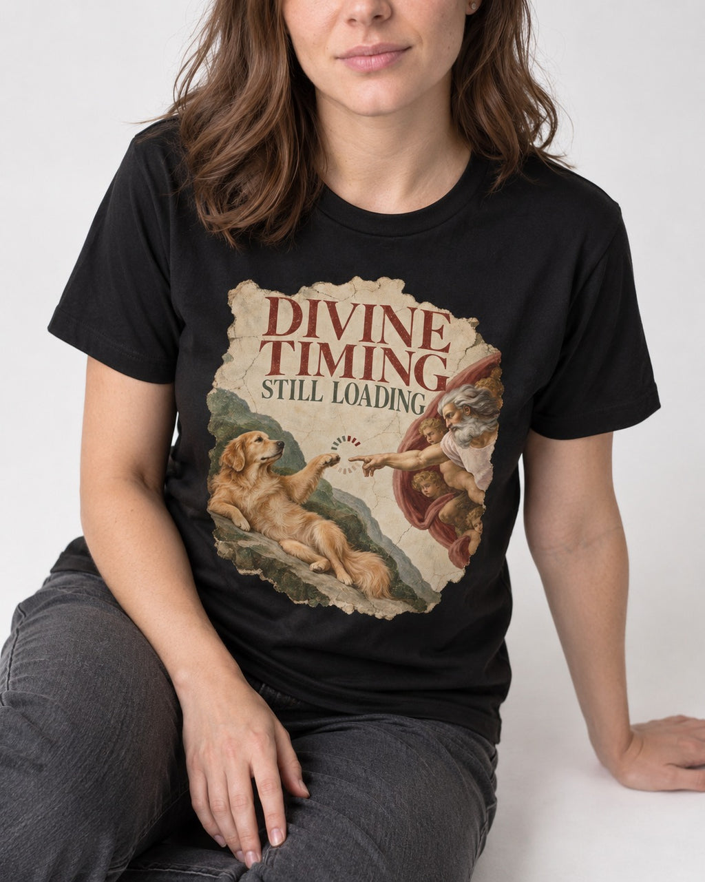 Divine Timing Still Loading Creation Scene Golden Retriever Graphic T-Shirt in Black, worn by a seated model