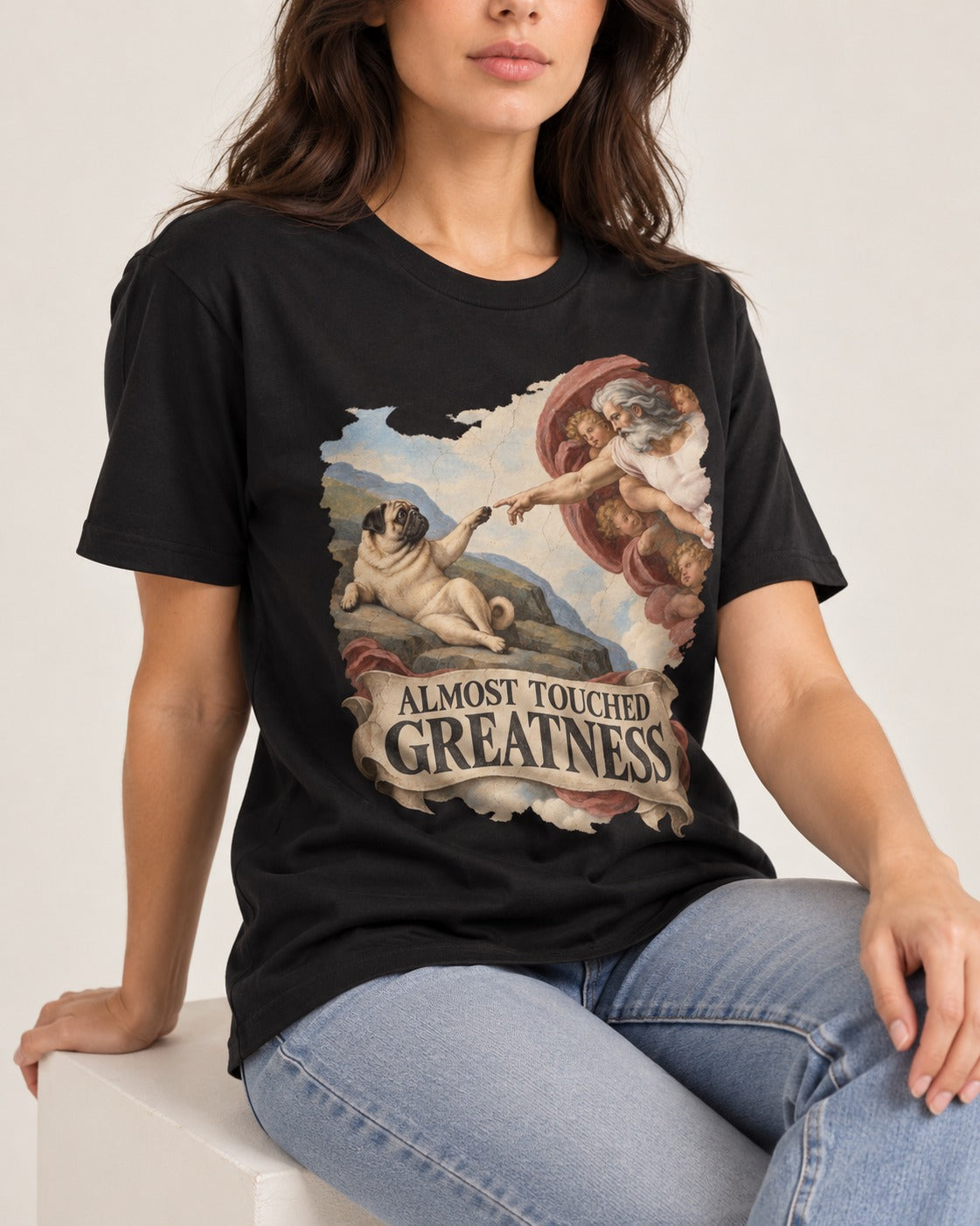 Pug Almost Touched Greatness Creation Parody Graphic T-Shirt in Black, worn by a seated model