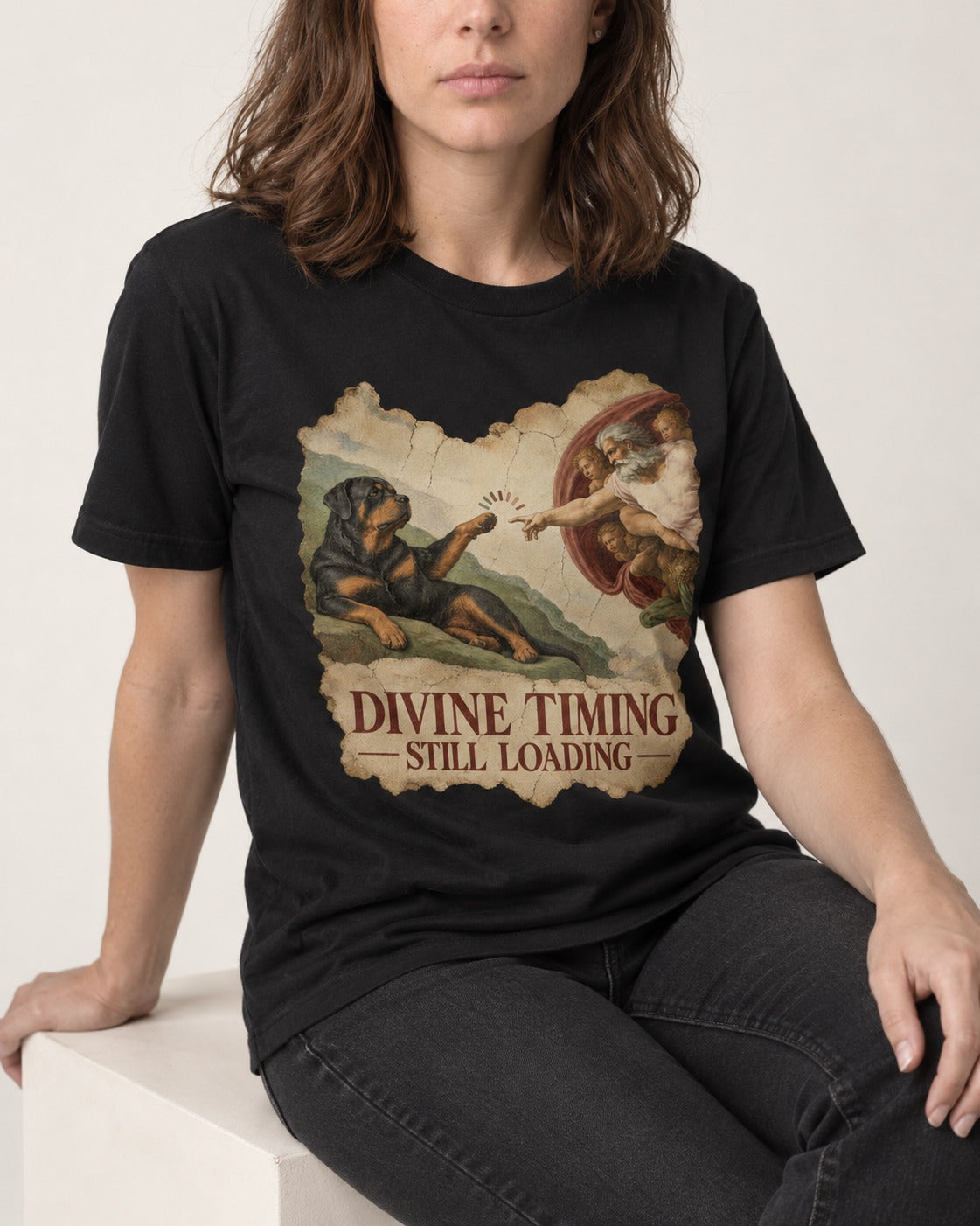 Rottweiler Divine Timing Still Loading Creation Parody Graphic T-Shirt in Black, worn by a seated model