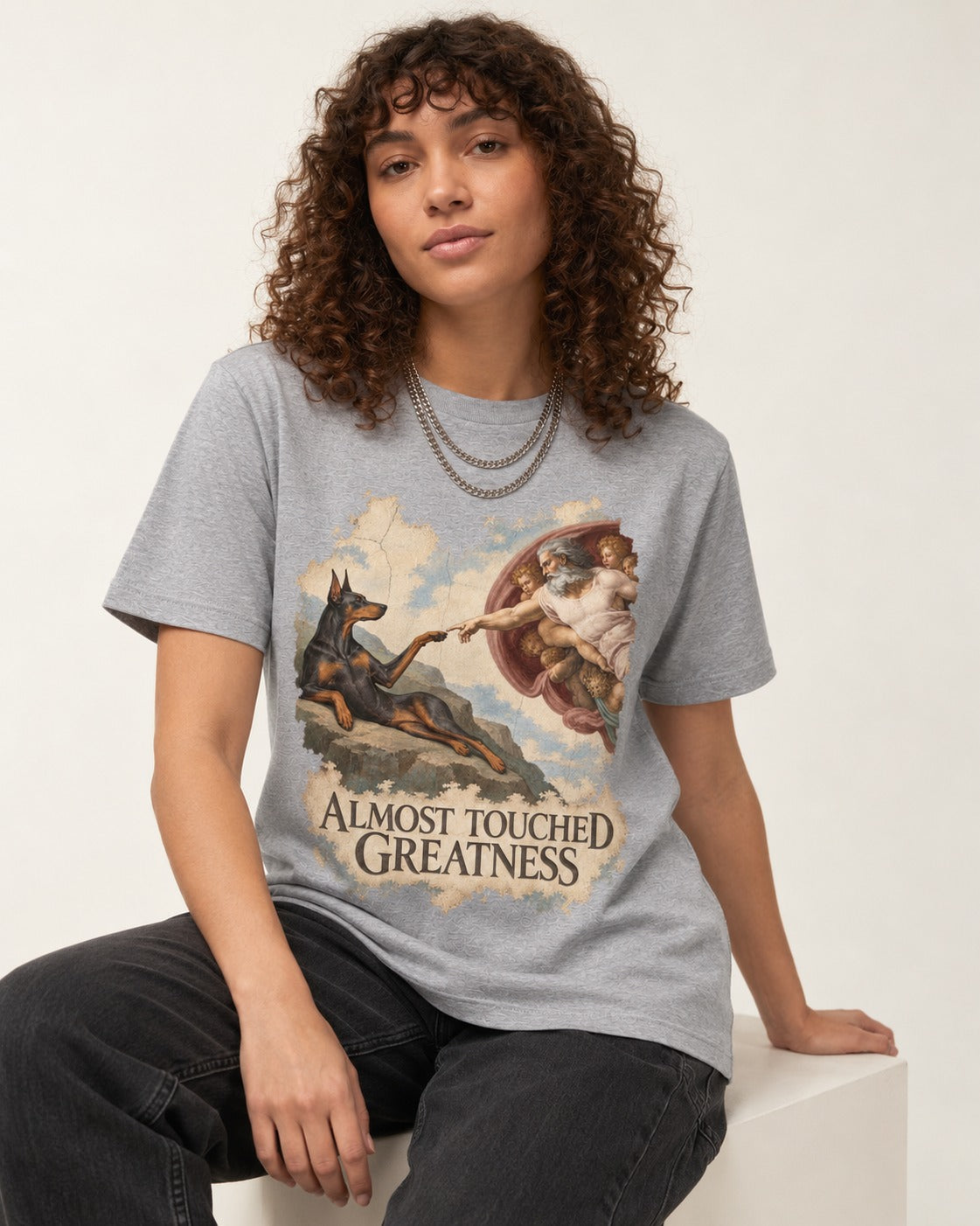 Doberman Almost Touched Greatness Creation Scene Graphic T-Shirt in Sport Grey, worn by a seated model