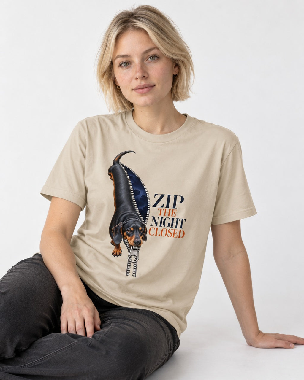 Zip the Night Closed Dachshund T-Shirt in Sand, worn by a seated model