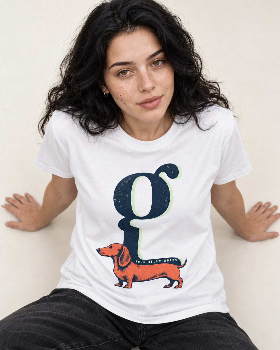 Dachshund Room Below Words Lowercase G Graphic T-Shirt in White, worn by a seated model