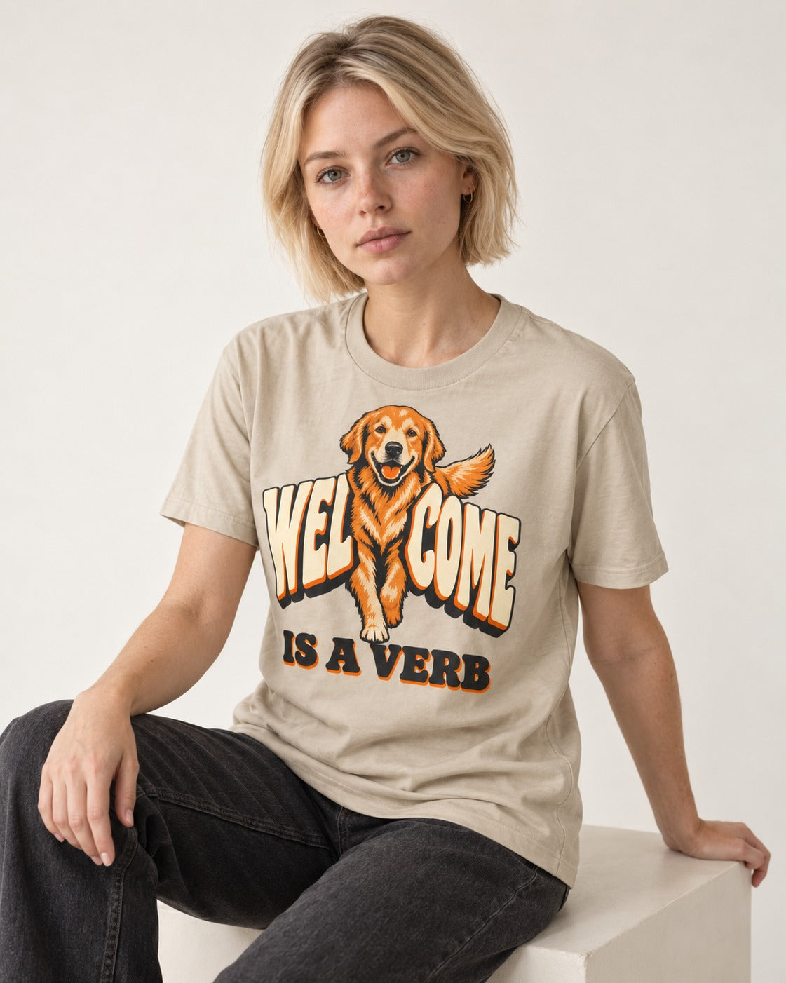 Golden Retriever Welcome Is a Verb T-Shirt in Sand, worn by a seated model