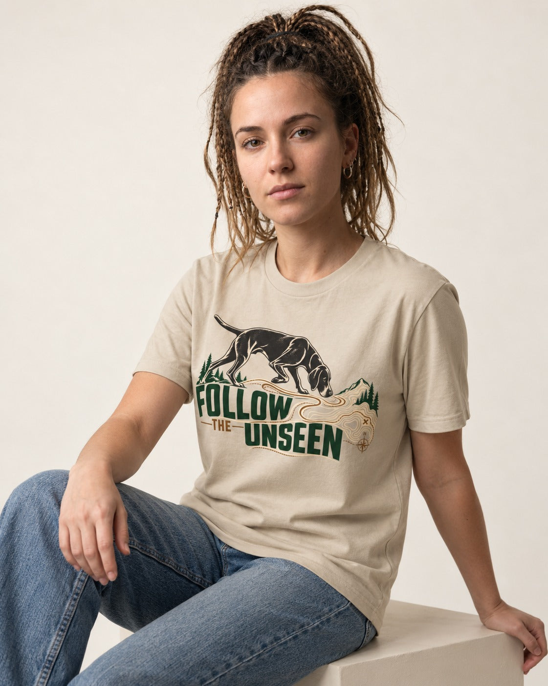 Follow the Unseen German Shorthaired Pointer T-Shirt in Sand, worn by a seated model