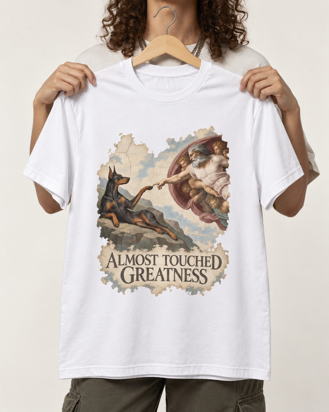 Doberman Almost Touched Greatness Creation Scene Graphic T-Shirt in White, held up on a hanger