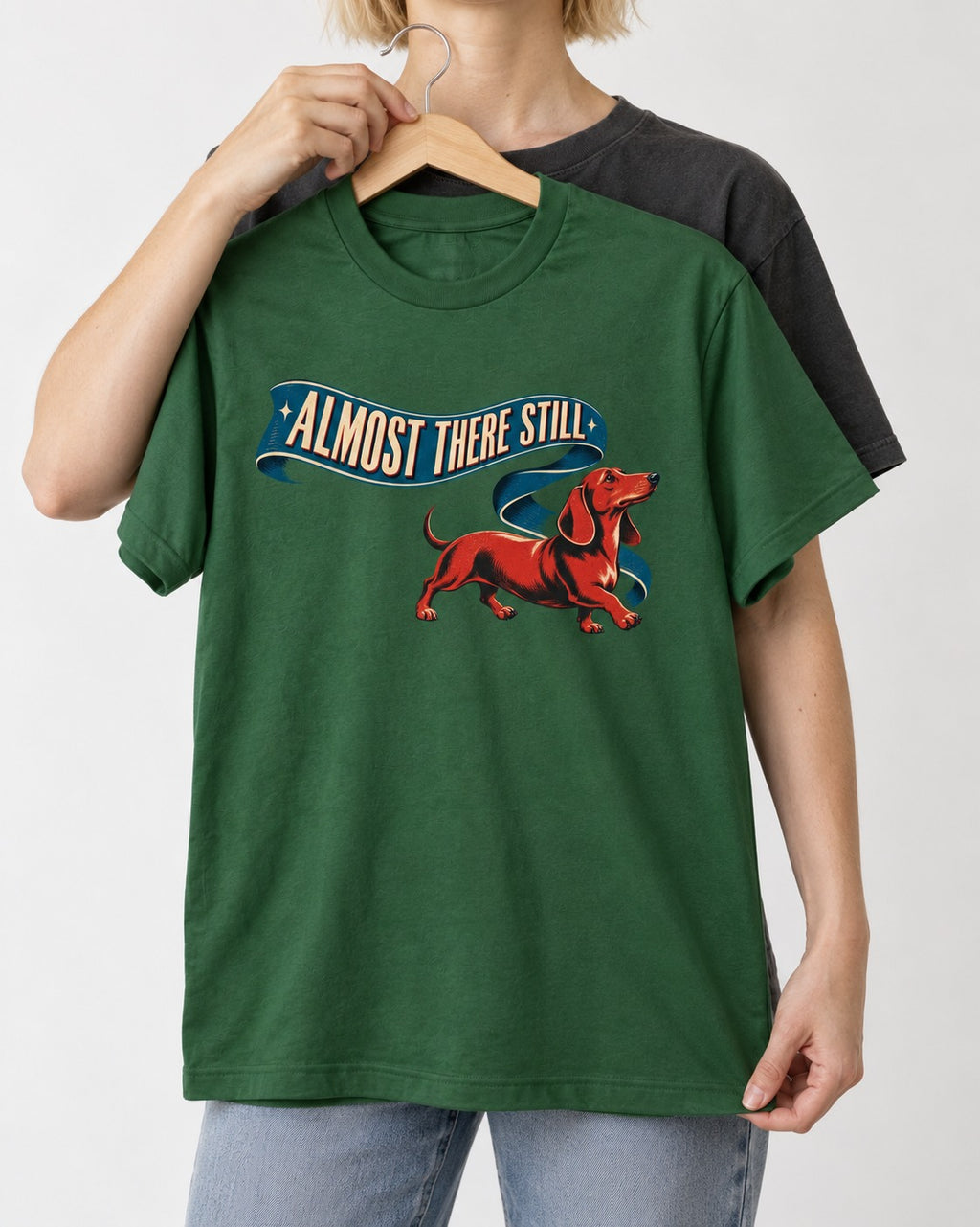 Dachshund Almost There Still Ribbon Graphic T-Shirt in Forest Green, held up on a hanger