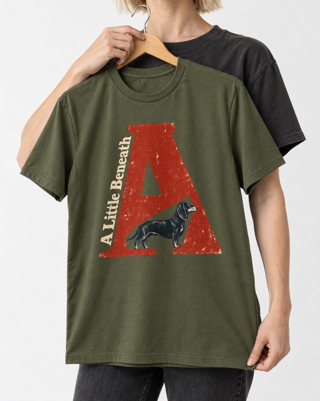 Dachshund A Little Beneath Monumental Letter Graphic T-Shirt in Heather Military Green, held up on a hanger
