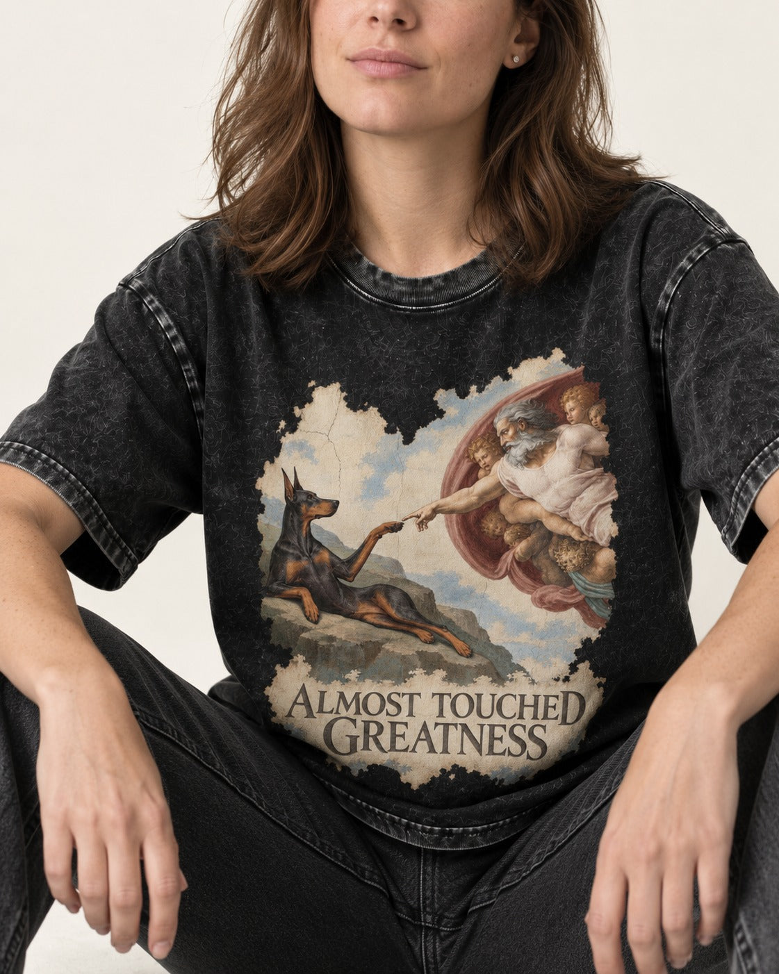 Doberman Almost Touched Greatness Creation Scene Graphic Washed Tee in Black, worn by a seated model
