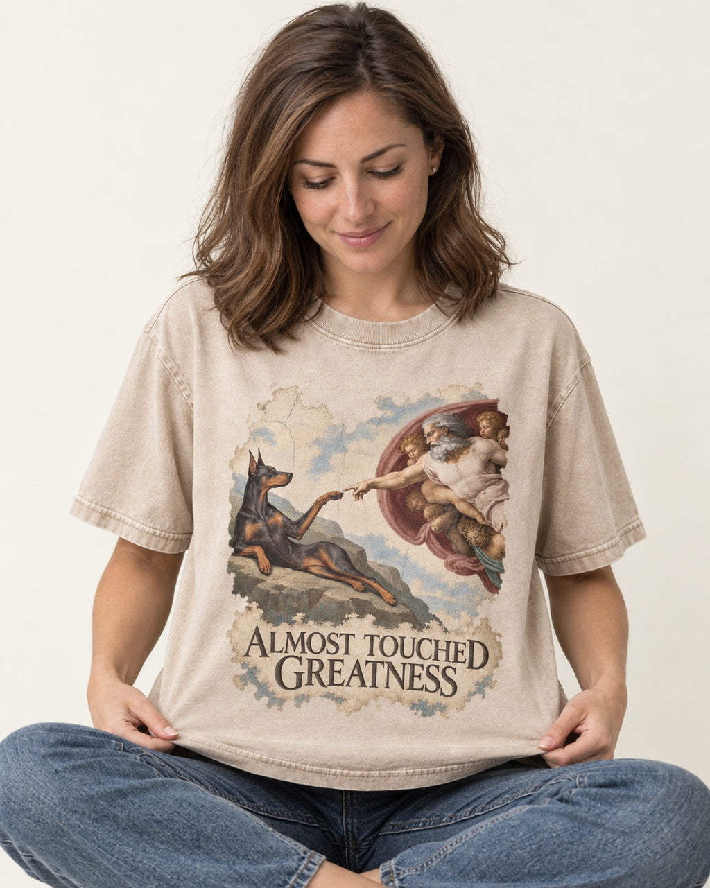 Doberman Almost Touched Greatness Creation Scene Graphic Washed Tee in Apricot, worn by a seated model