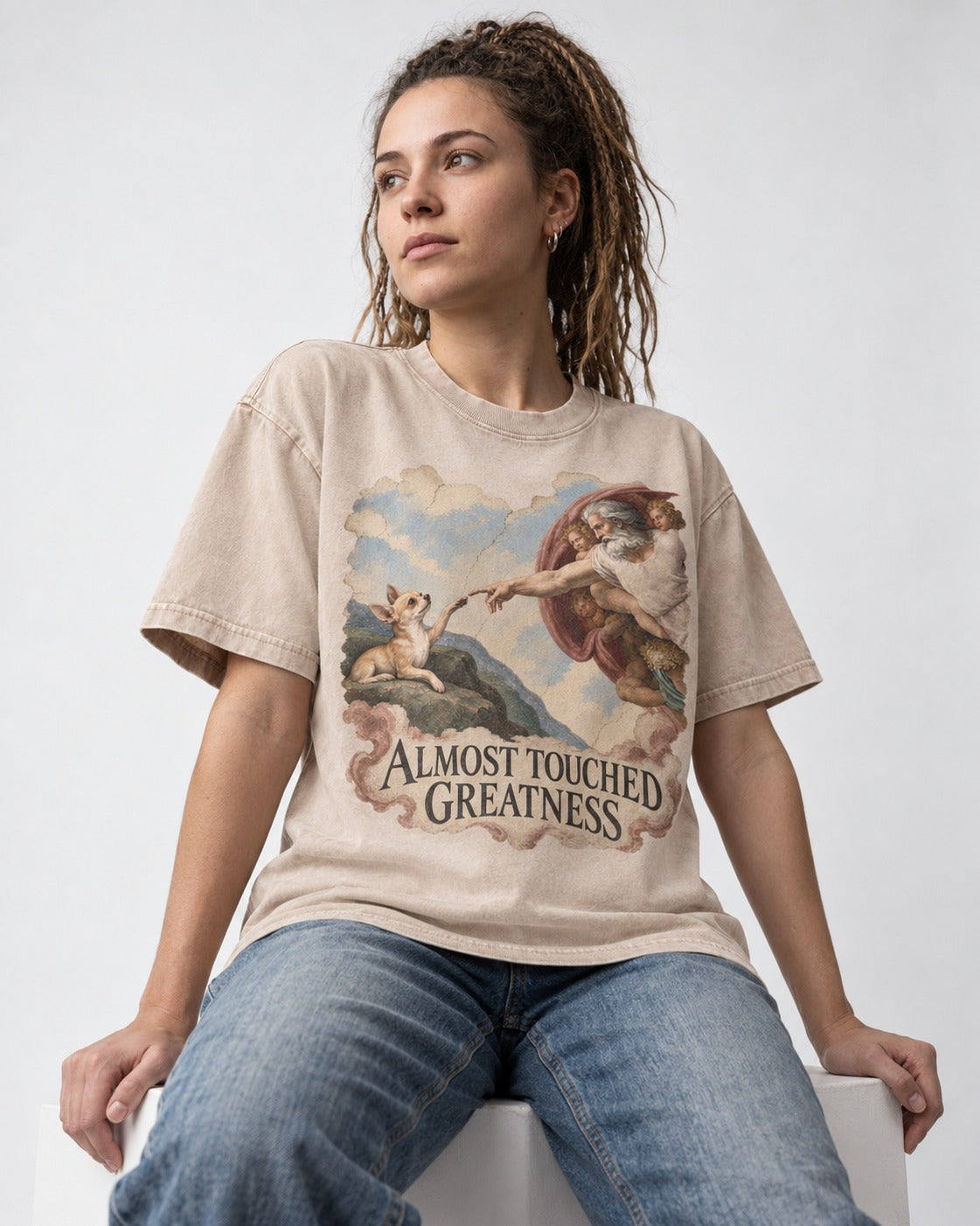Chihuahua Almost Touched Greatness Renaissance Parody Washed Tee in Apricot, worn by a seated model