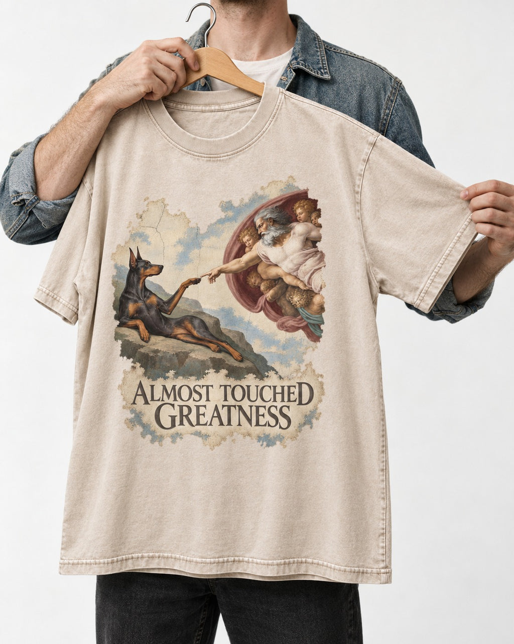 Doberman Almost Touched Greatness Creation Scene Graphic Washed Tee in Apricot, held up on a hanger